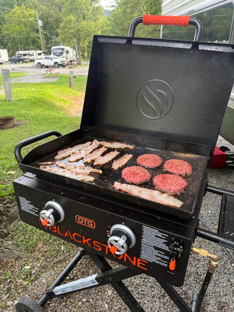 Bacon and seasoned burger patties cooking on our Blackstone On the Go griddle beside the camper