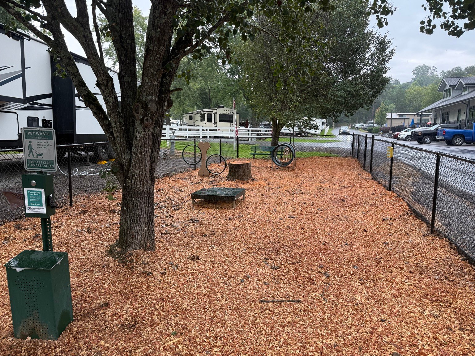 Fenced dog park at Clabough's Campground with shade trees, wood chips, hoops, a platform, and a pet-waste station.