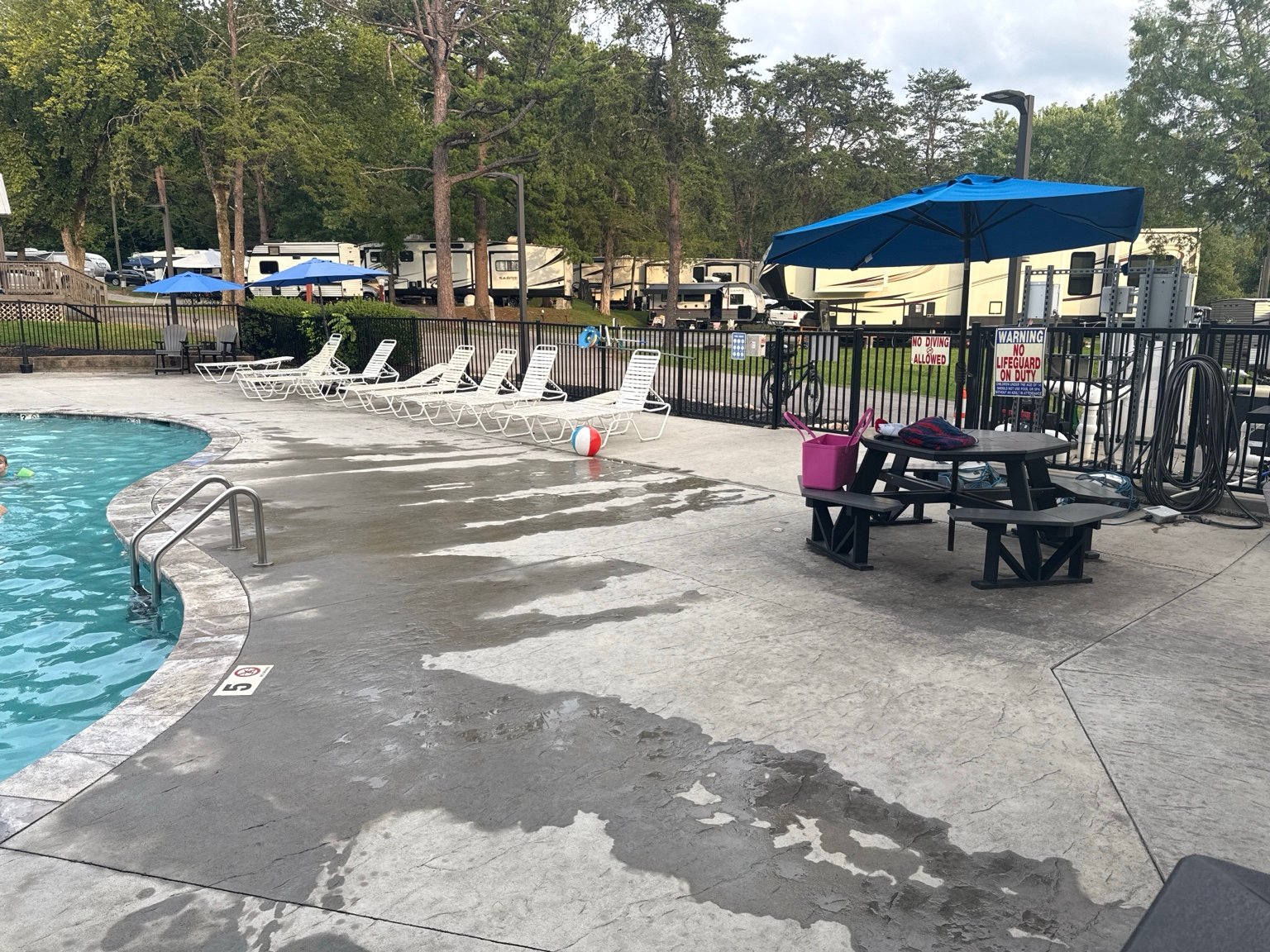 Main pool seating at Clabough's Campground with lounge chairs, blue umbrellas, picnic tables, and RV sites nearby.